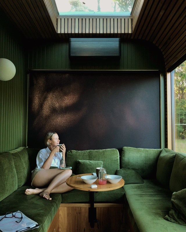 Woman sitting on green velvet couch, sipping from a cup, inside a green-walled room with a small round table.