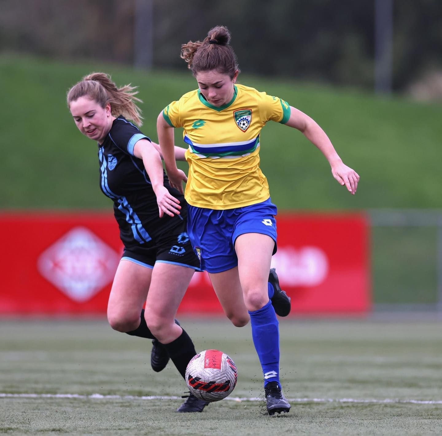 Two women are playing soccer and one is wearing a yellow shirt