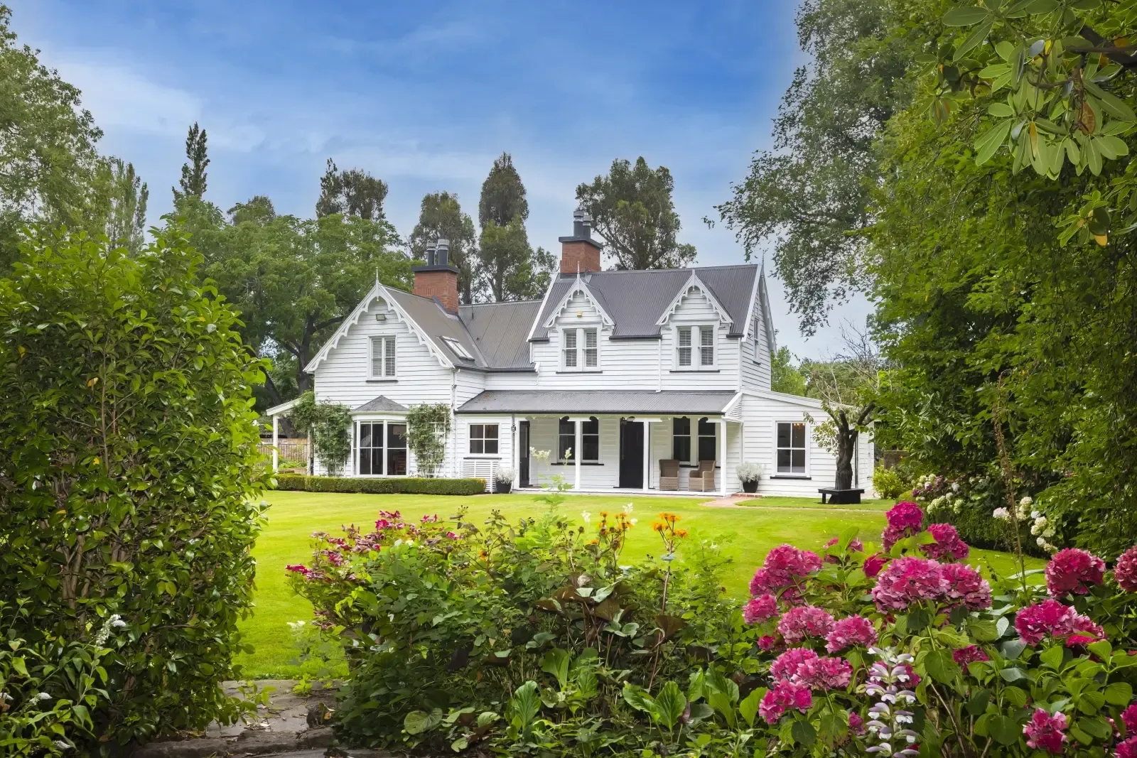 A large white house is surrounded by bushes and flowers.