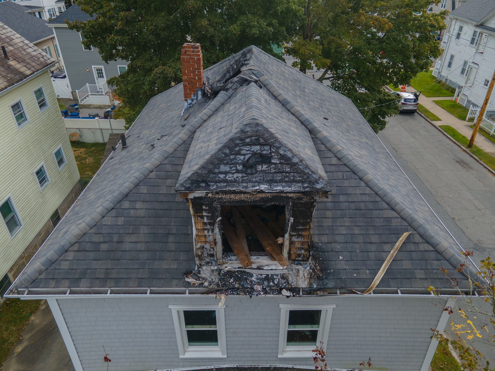 Damaged house roof with a burnt-out dormer and chimney. Gray shingles, wooden structure, street view.