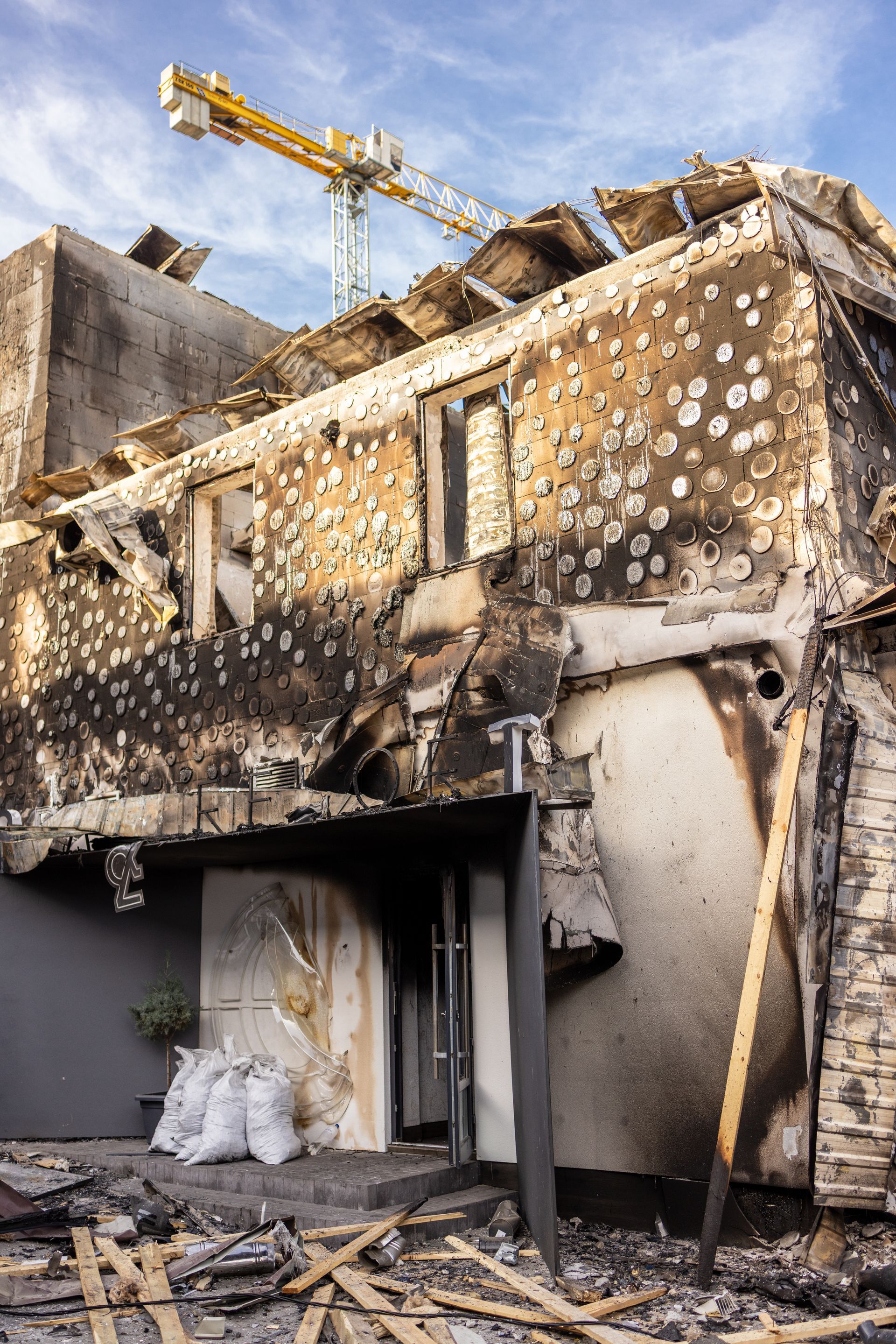 Burnt building facade, riddled with holes, damaged roof, construction crane in the background, blue sky.