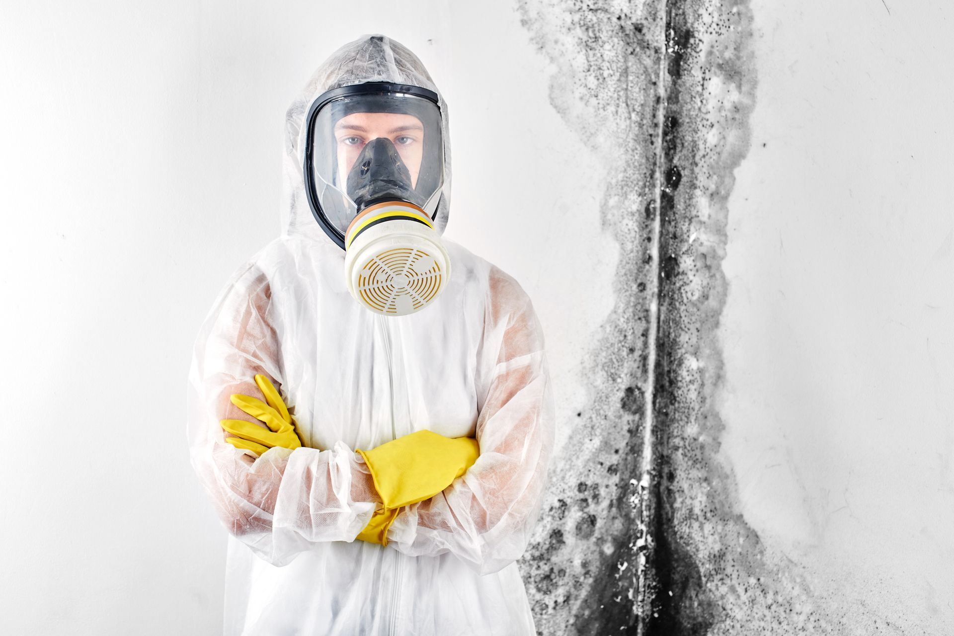 Person in protective suit and respirator mask standing in front of wall with mold.