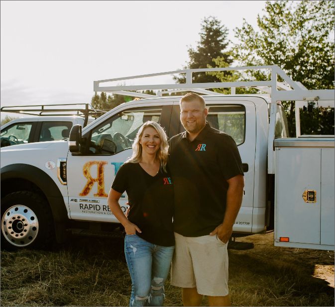 Man and woman stand by a white work truck with logo, smiling. Outdoors, sunny.