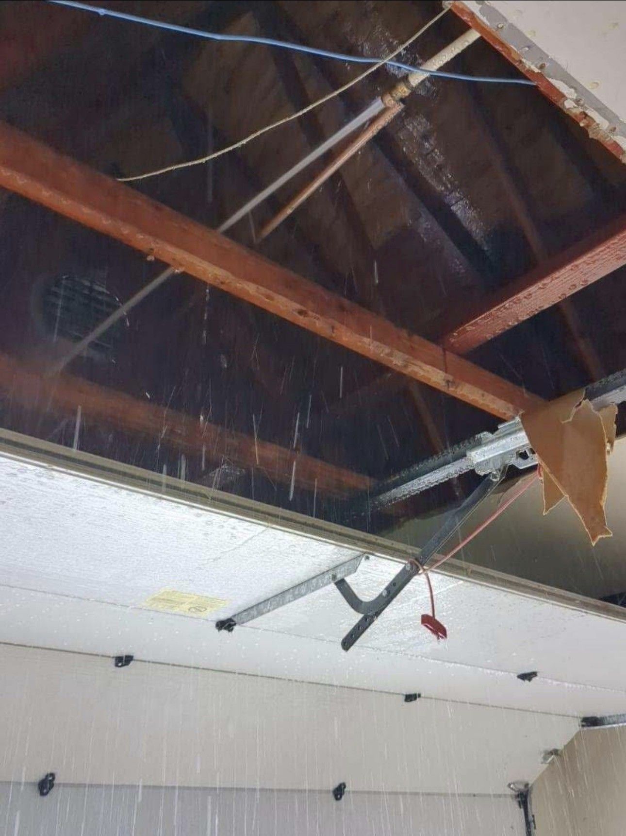 Water leaking from a damaged garage ceiling, brown wooden beams, a broken overhead door track, and exposed wiring.