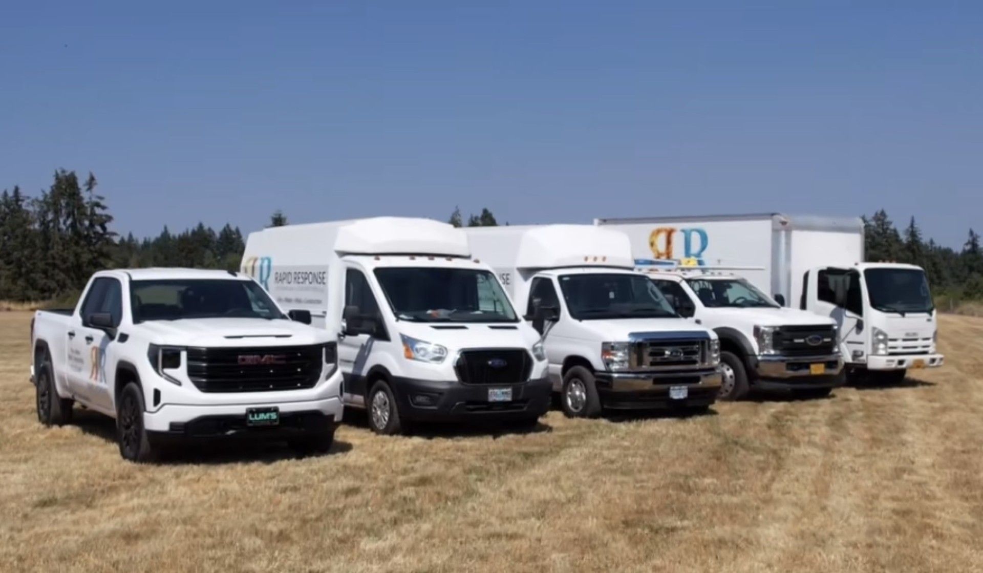 Five white commercial vehicles parked on dry grass under a blue sky. The vehicles have company logos.