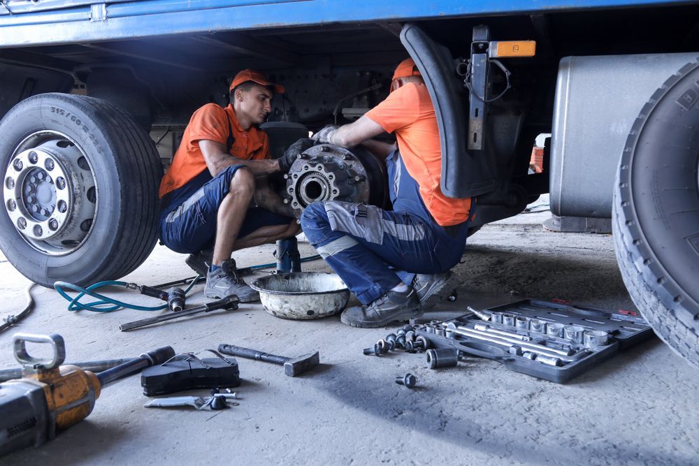 Two Mechanics Repair a Truck Wheel Under a Lifted Vehicle in a Garage — Forster Smash Repairs in Forster, NSW