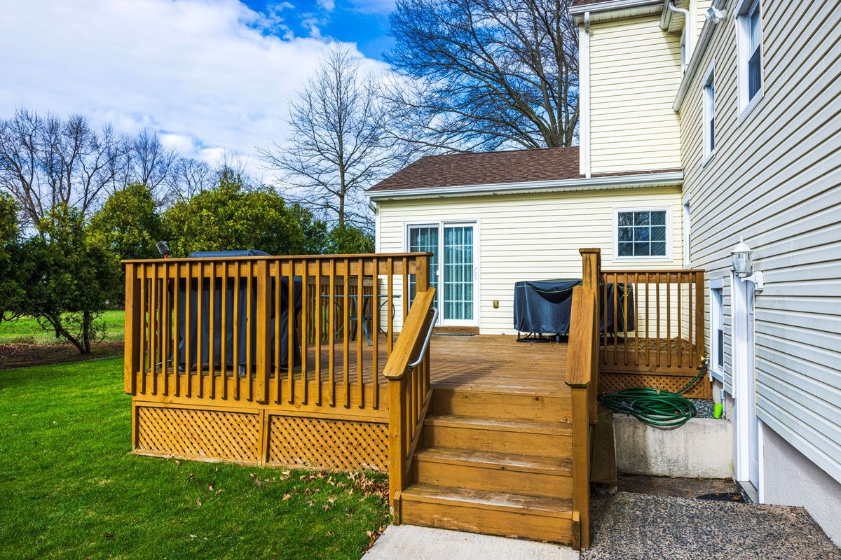Wooden deck with stairs, leading to a house with a sliding glass door. Green lawn and trees in background.