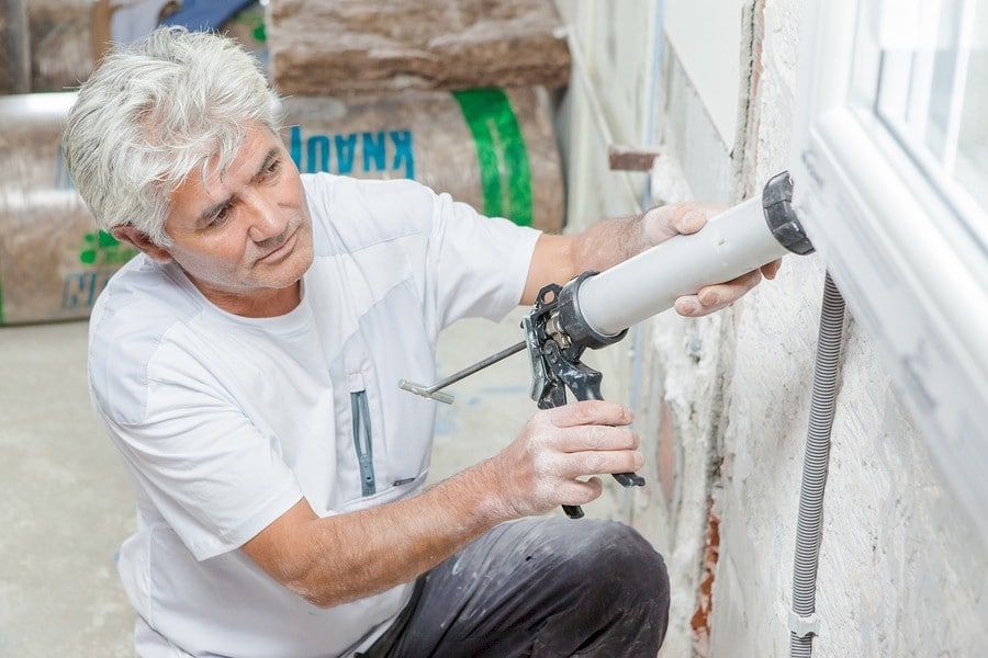 Man caulking around a window. He wears a white shirt, gray hair, and is indoors with insulation