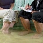 Two people in a flooded room, sitting on a sofa while a person with a pen in hand takes notes on a clipboard. Two people in a flooded room, sitting on a sofa while a person with a pen in hand takes notes on a clipboard.