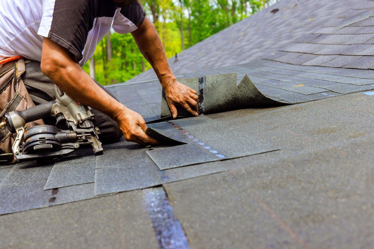 Roofer installing dark gray asphalt shingles on a roof, with tools visible; sunny setting.