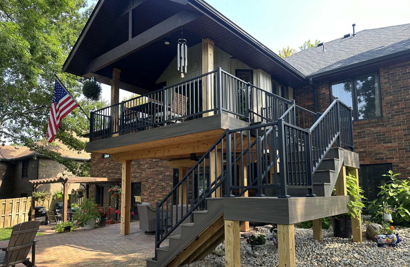 Deck under construction; wooden frame with installed gray deck boards and vertical posts, set against a light blue house.