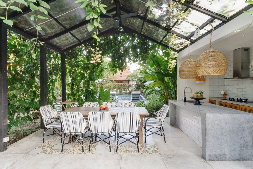 Outdoor dining area with a table, chairs, and kitchen under a glass roof, surrounded by plants.