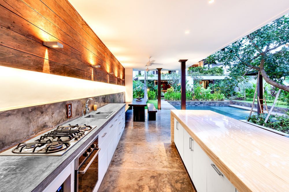 Modern kitchen with wood, concrete, and white cabinetry, opening to a lush outdoor area.