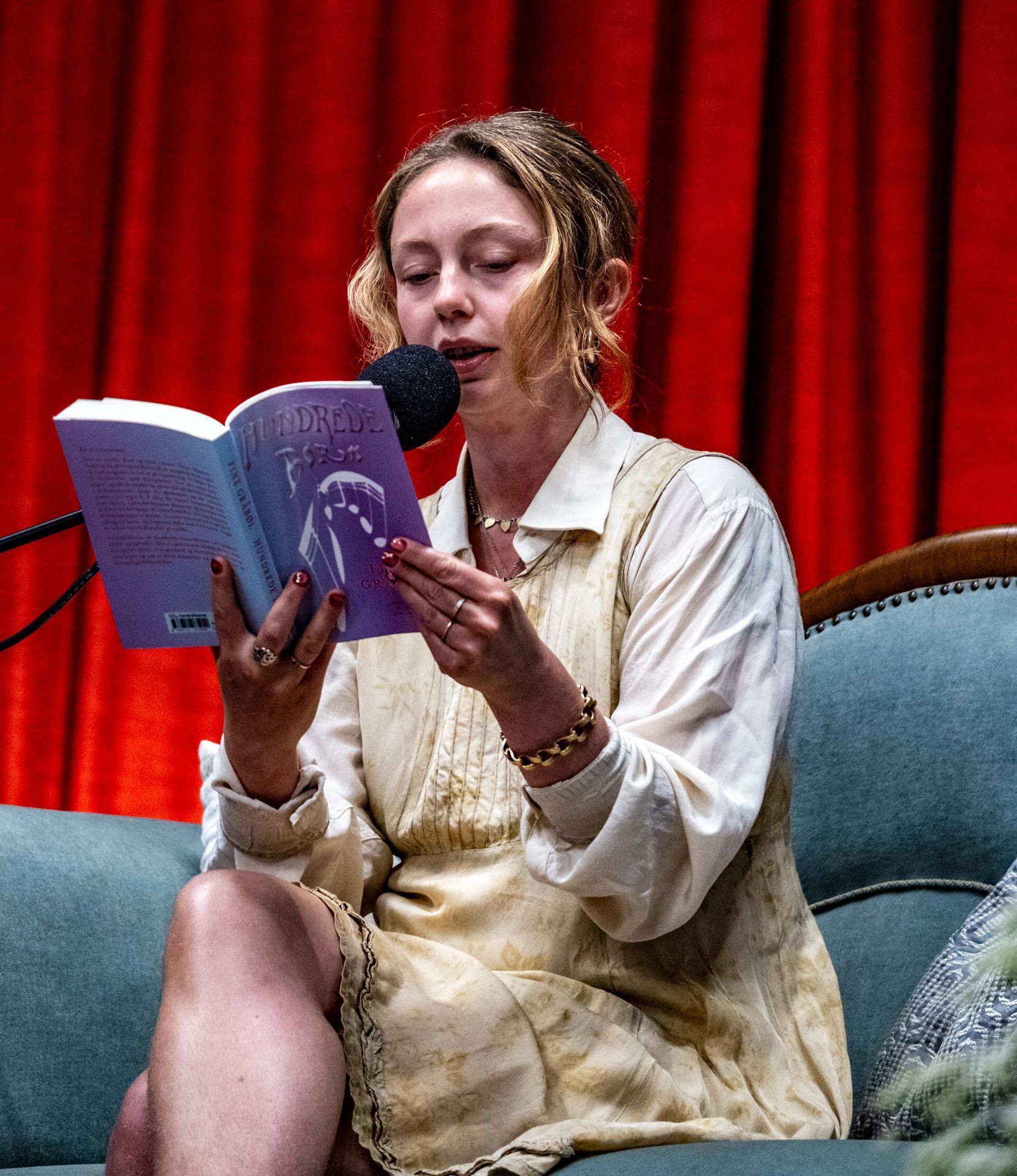 En kvinde sidder på en sofa og læser en bog ind i en mikrofon.