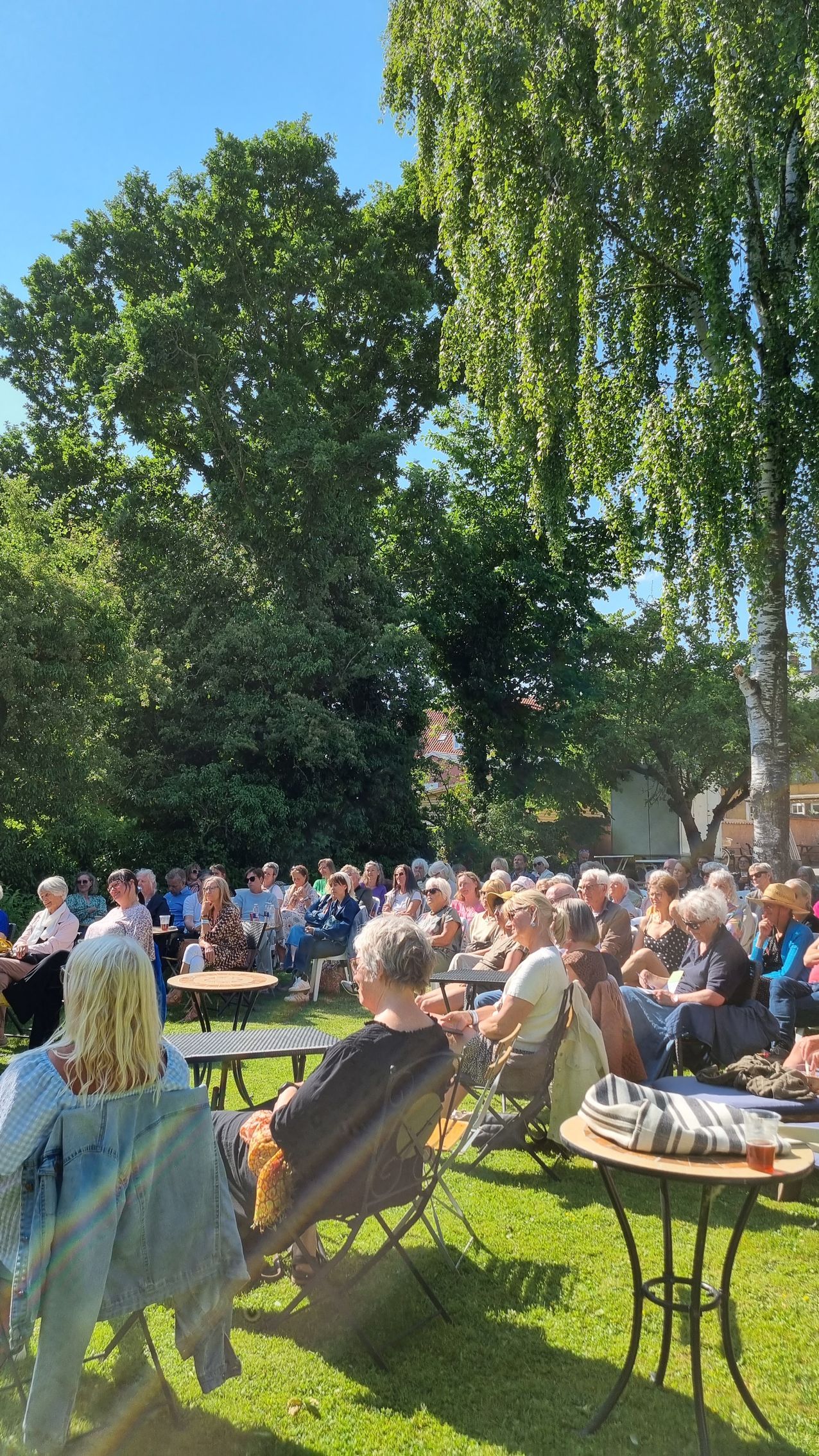 En gruppe mennesker sidder ved borde i en park.