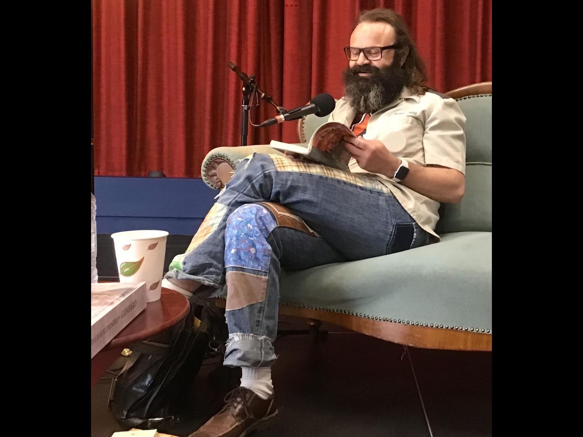 En mand med skæg sidder på en sofa og læser en bog.