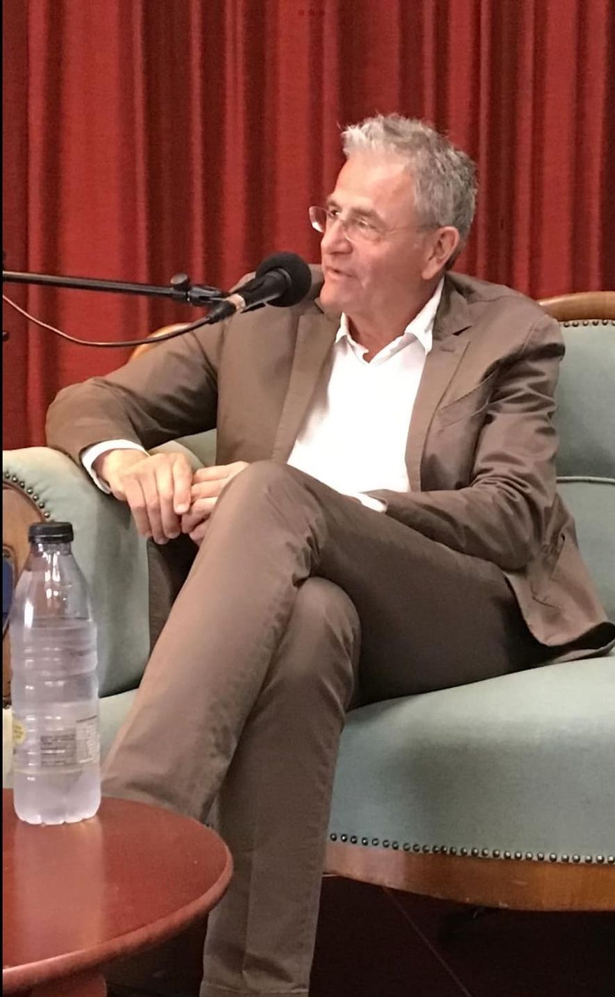 En mand i jakkesæt sidder på en sofa foran en mikrofon.