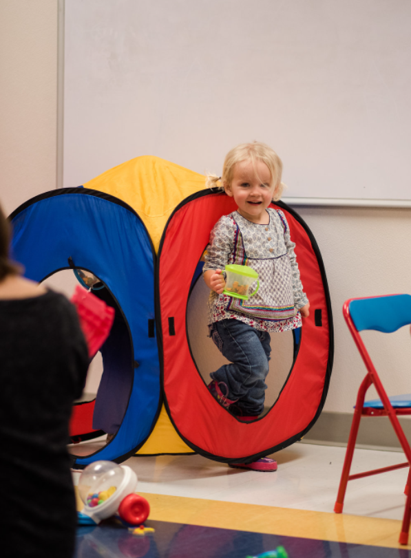 A little girl is standing in a colorful tent holding a toy.
