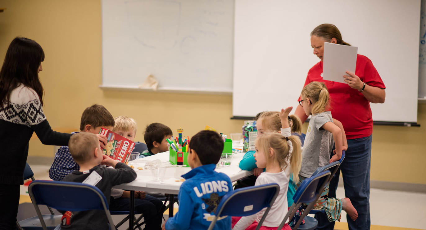 A group of children are sitting around a table with a woman standing behind them.