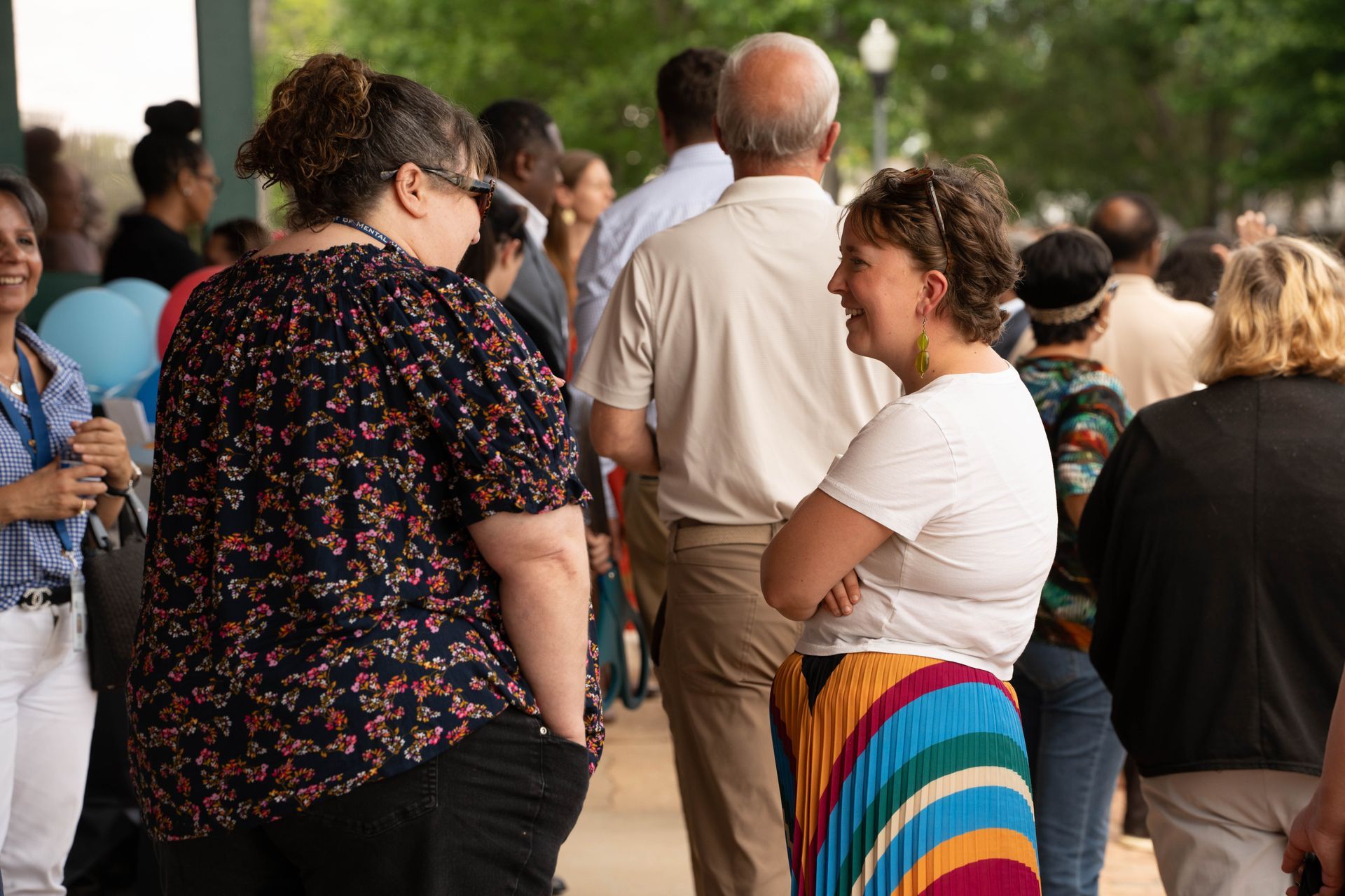 People gathered outside, conversing. A woman in a floral top talks to another in a white tee and rainbow skirt.