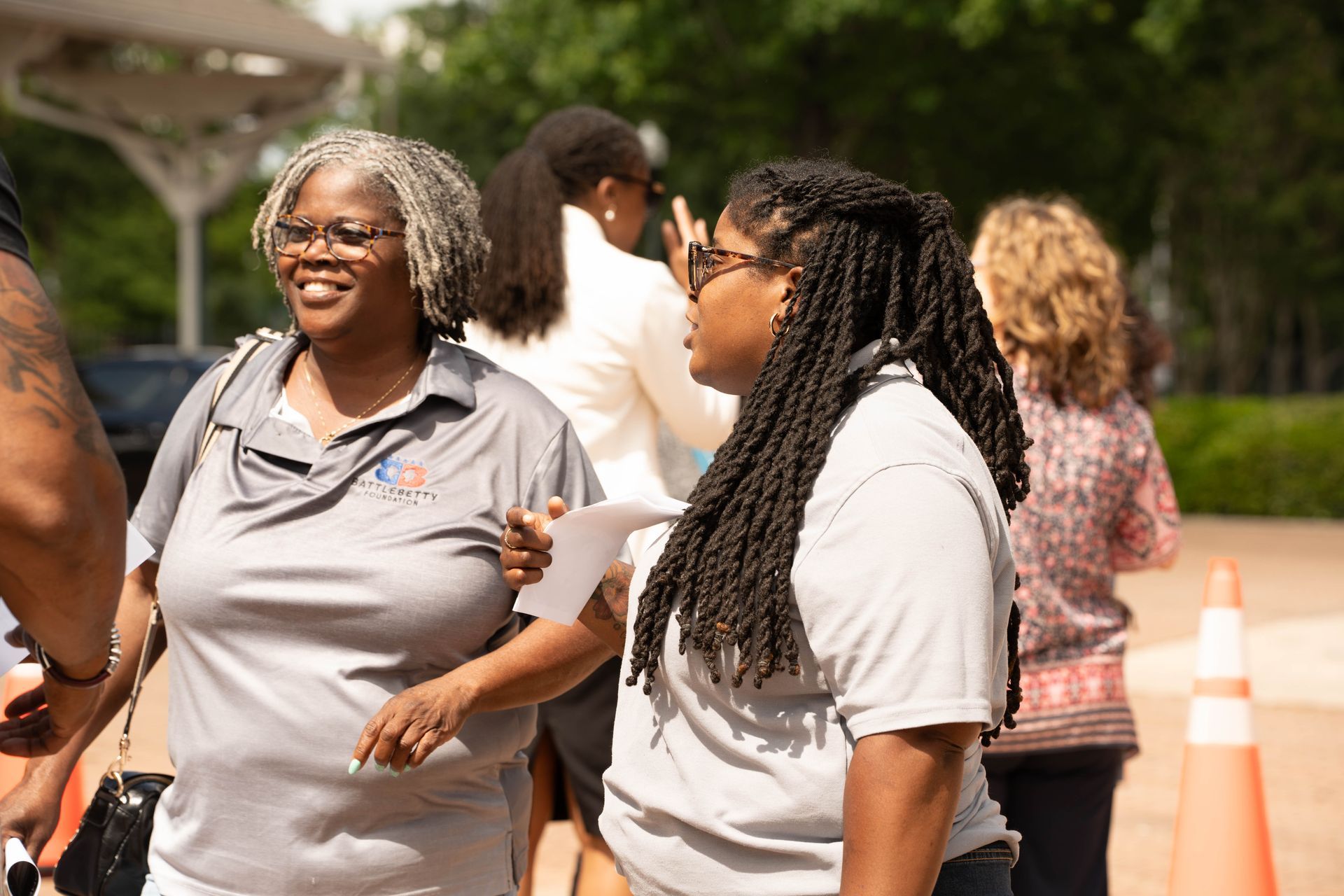 Two women smile and talk outdoors. One has gray hair, the other has dark braids. Other people are in the background.