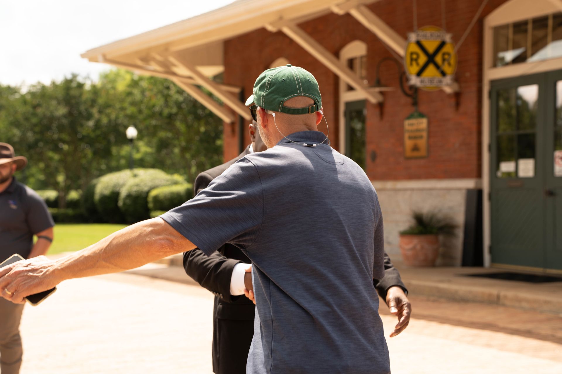 Man in green cap greets another man in front of a brick building with an 