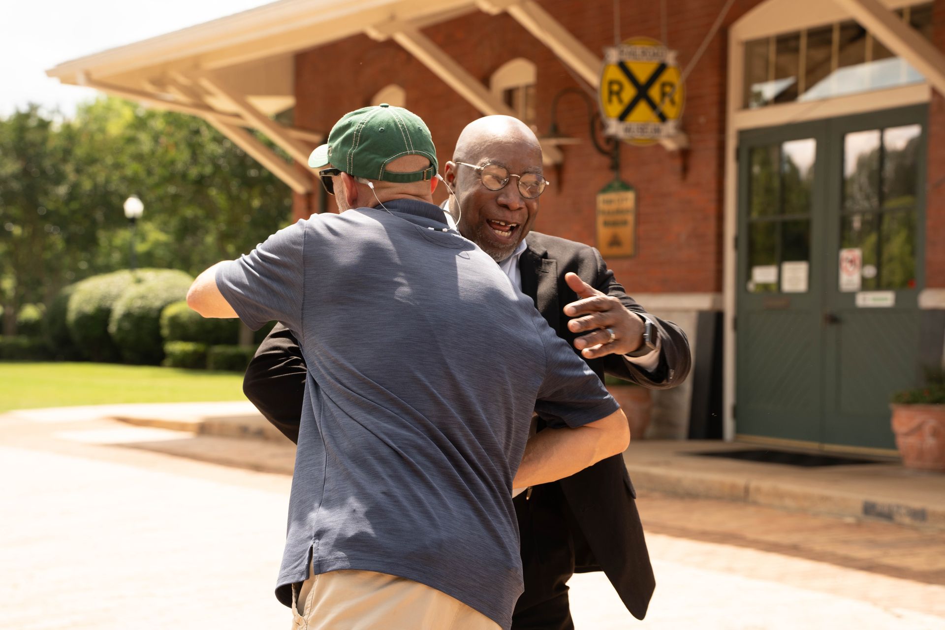 Two men embrace warmly outside a brick building with a RR sign.