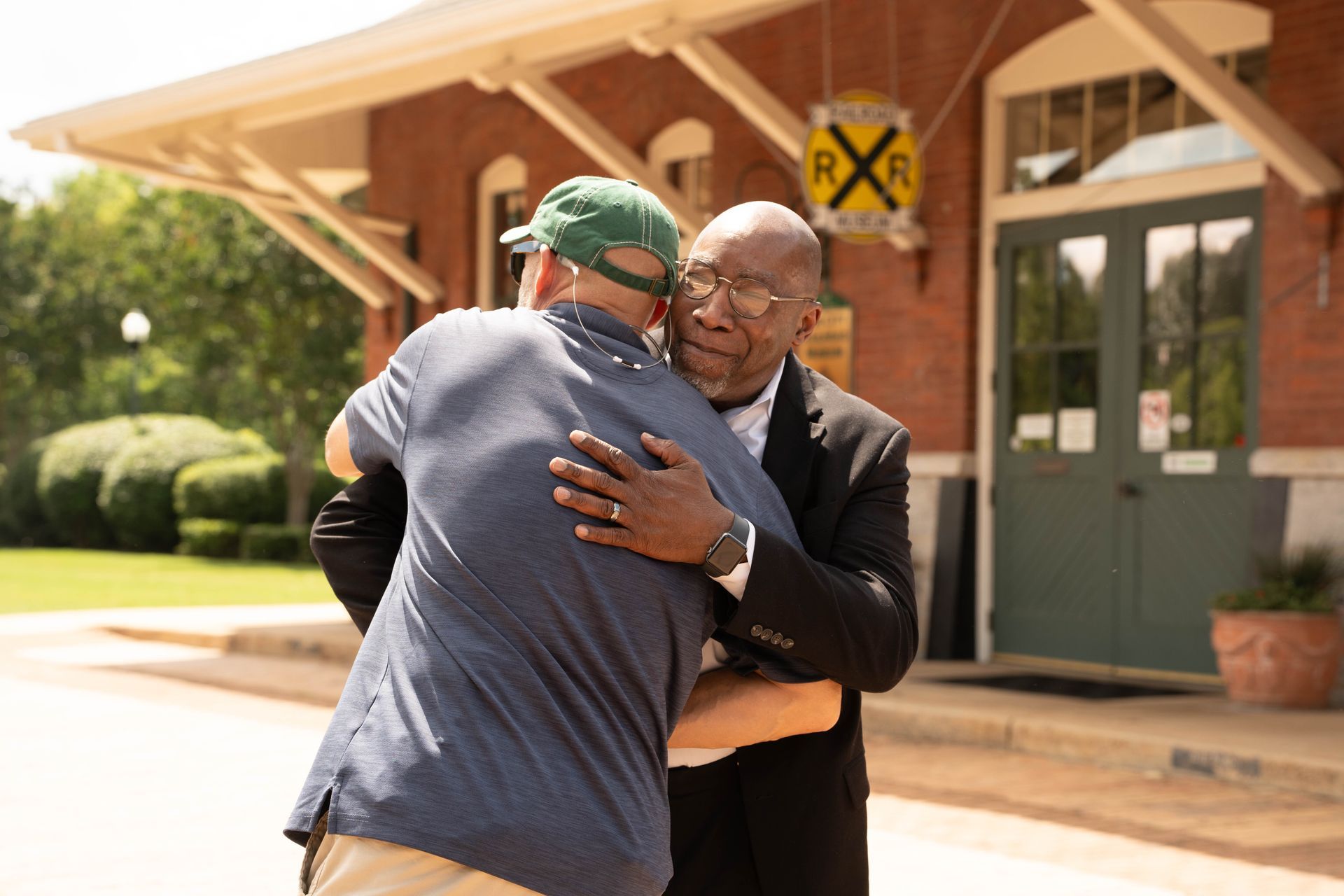 Two men embracing warmly outside a brick building with a railroad sign.