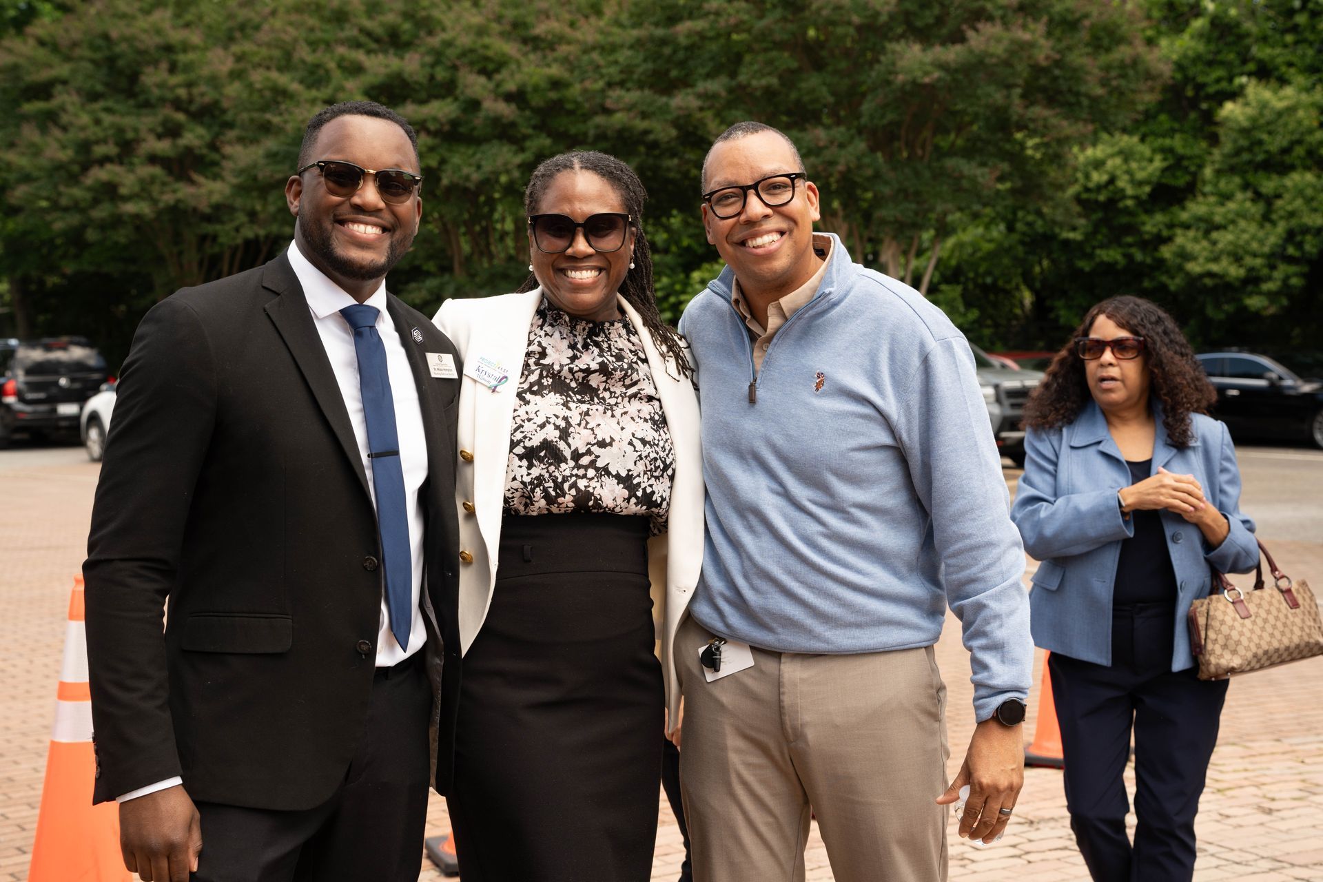 Three people smiling outdoors, two are embracing. Another woman is in the background.