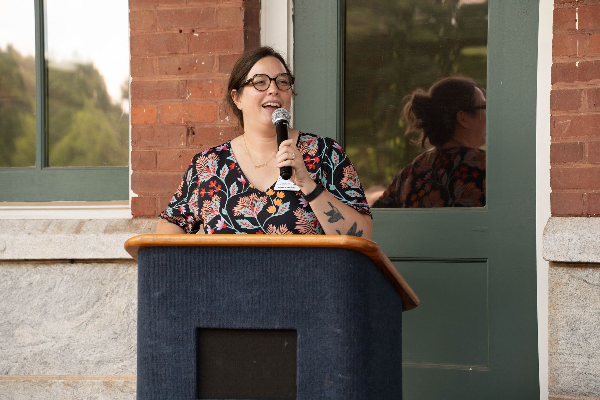 Woman with glasses speaking at a podium, smiling, outdoors in front of a brick building.