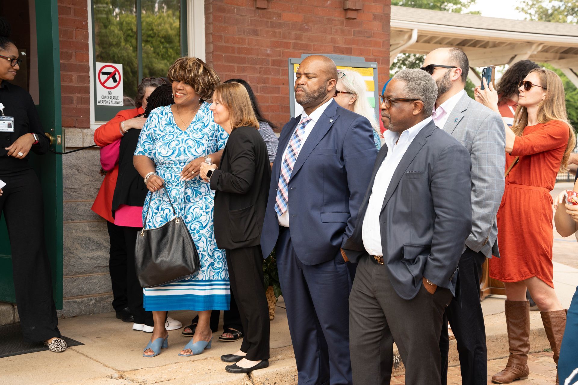Group of people outside brick building; man in blue suit and others look towards entrance.