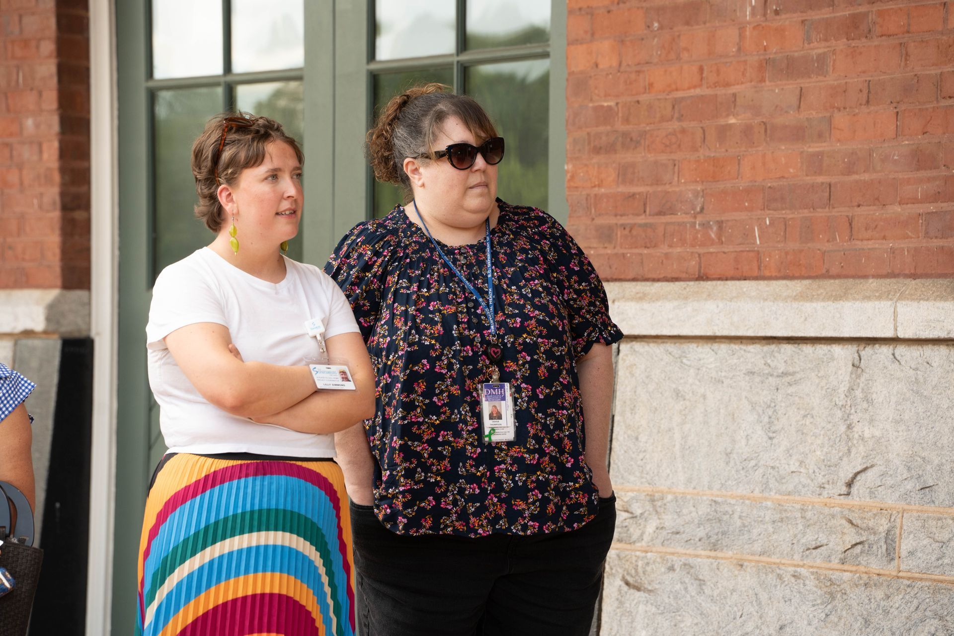 Two women stand outside a brick building. One wears a white shirt and rainbow skirt, the other a floral shirt, black pants, and sunglasses.