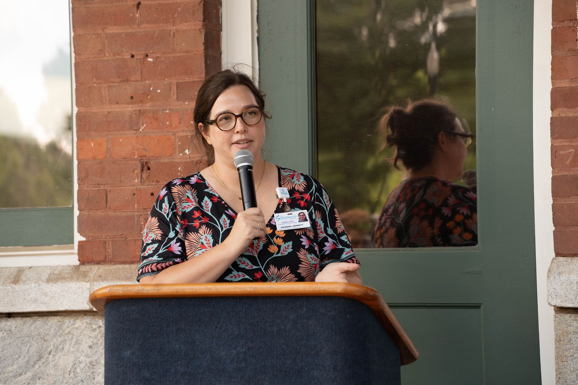 Woman with glasses speaks at a podium, outdoors in front of a brick building.