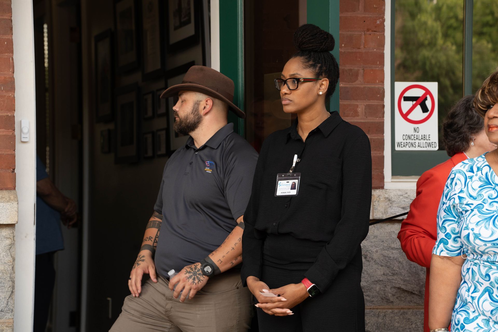 Man in hat and woman with bun stand outside, looking away. 