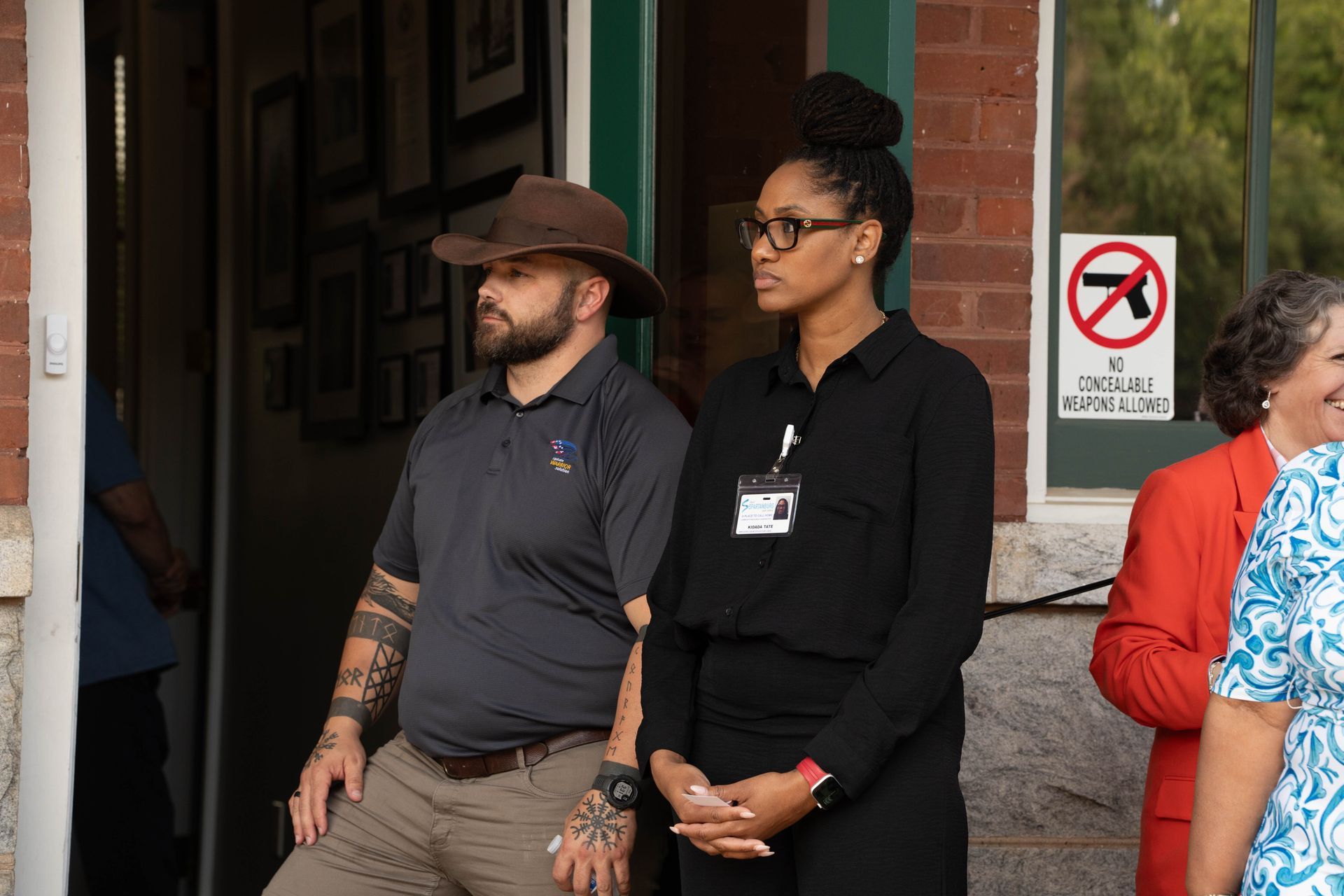 Man in hat and woman in black suit stand outdoors, watching something. 