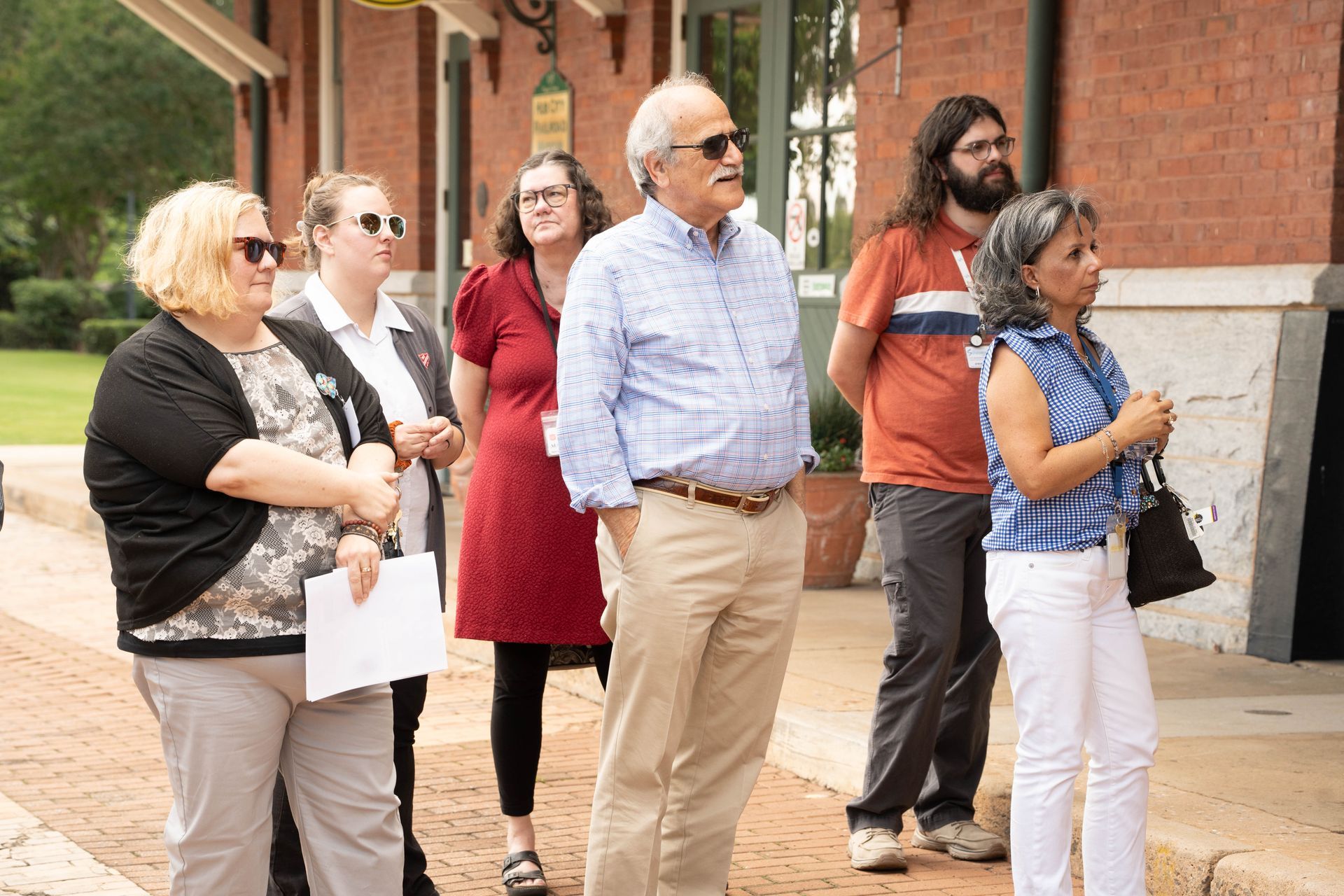 Group of people standing outside a brick building, observing.  Sunny day.