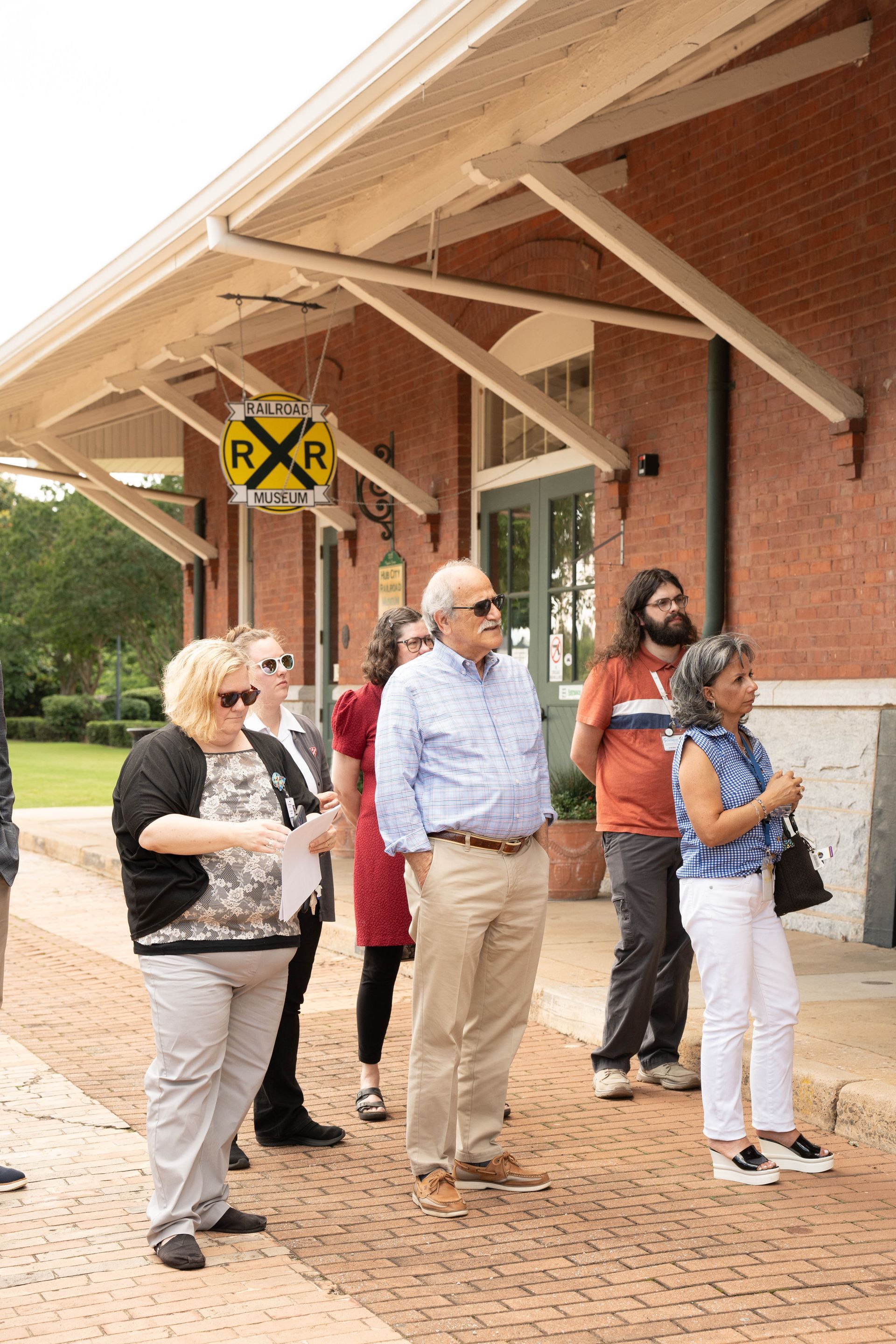 Group of people outside a brick building with a train crossing sign; some are looking upward.