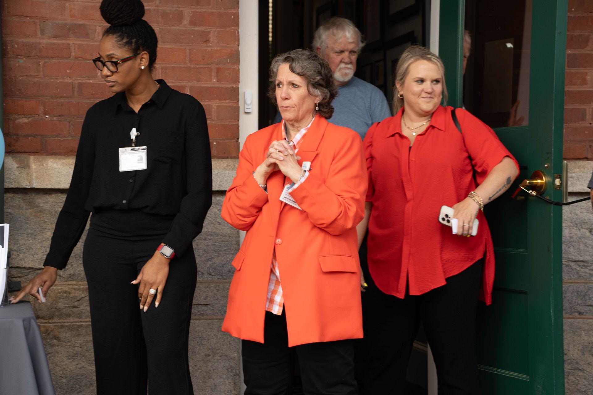 Four people stand in front of a building. Woman in orange jacket claps hands. Woman in red shirt holds phone.