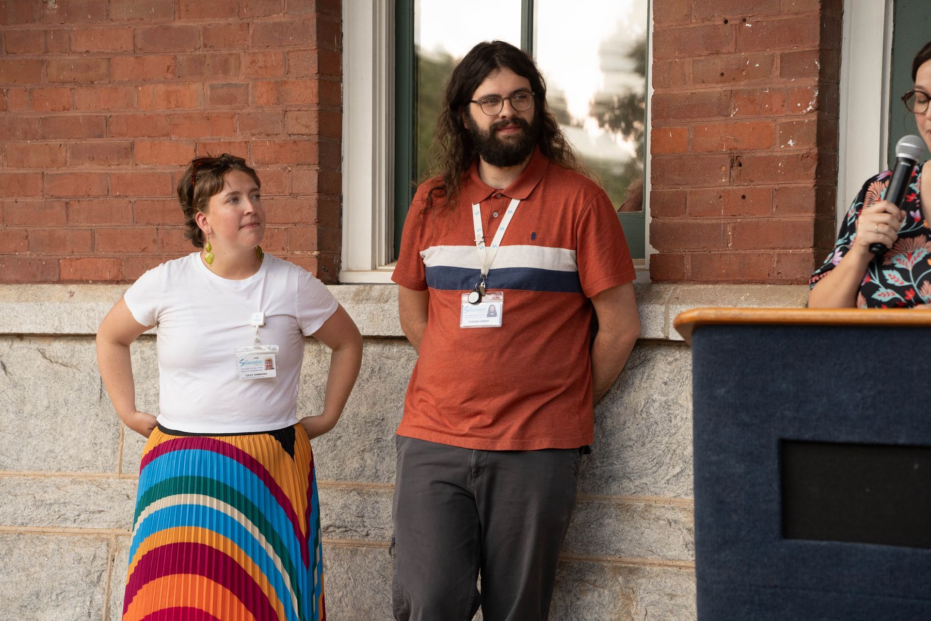 Three people stand outside near a brick building. Man with beard and glasses. Woman in rainbow skirt and white shirt. Person with microphone at podium.