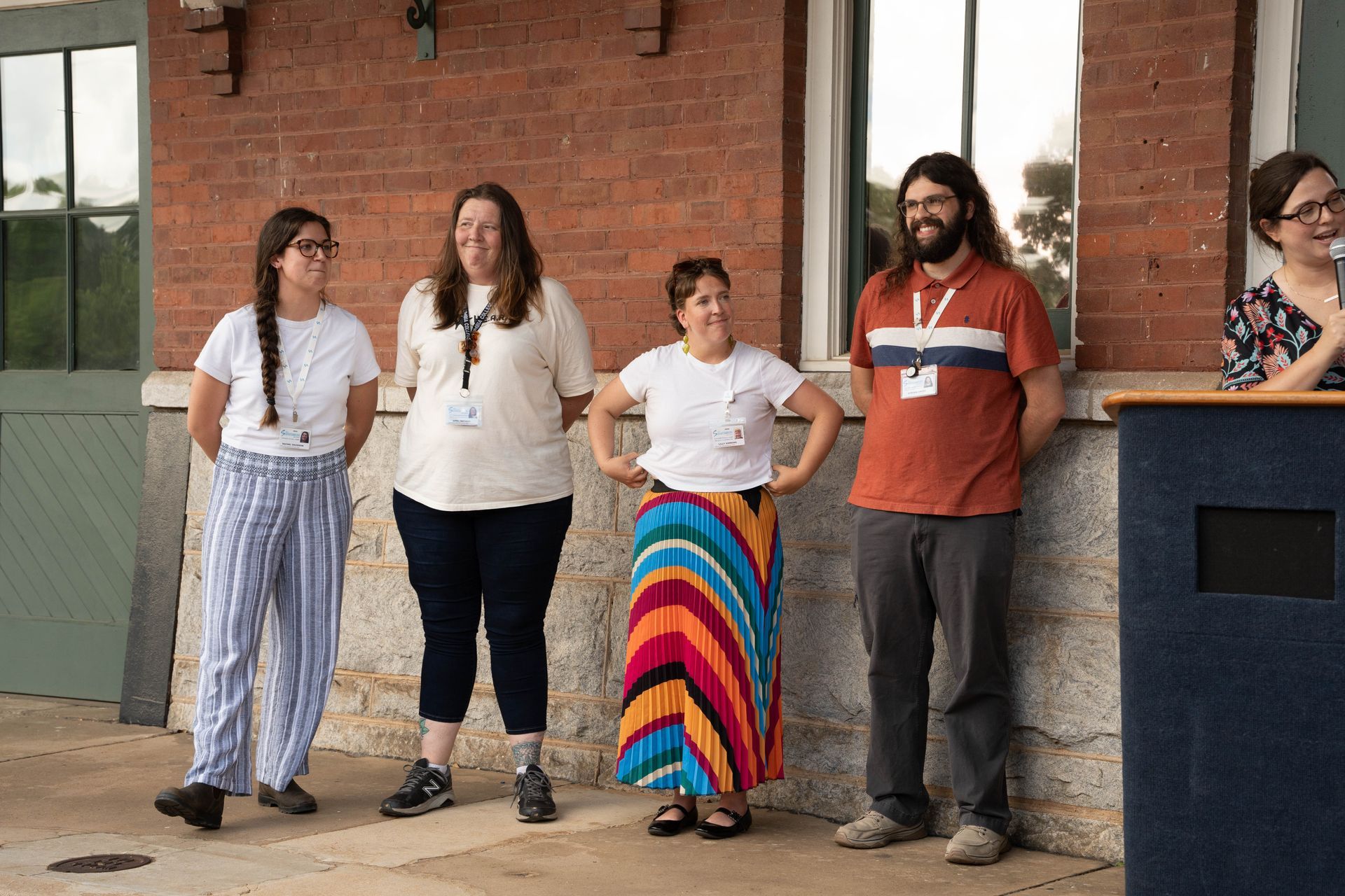 Five people stand outside a brick building. Three women and one man are smiling, with name tags. One woman is seated.