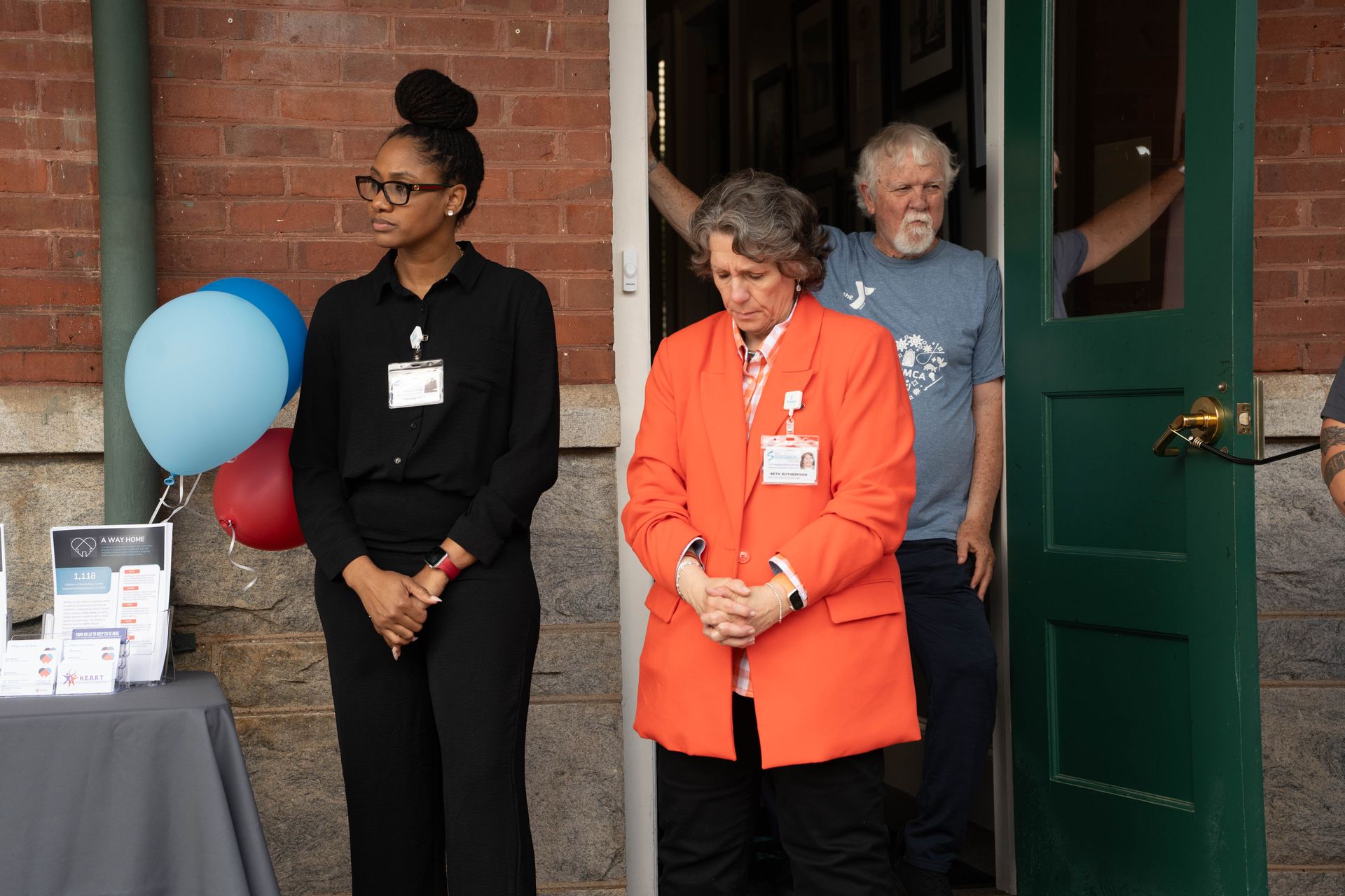 Three people standing in front of a building: a Black woman, a woman in orange, and a white man.