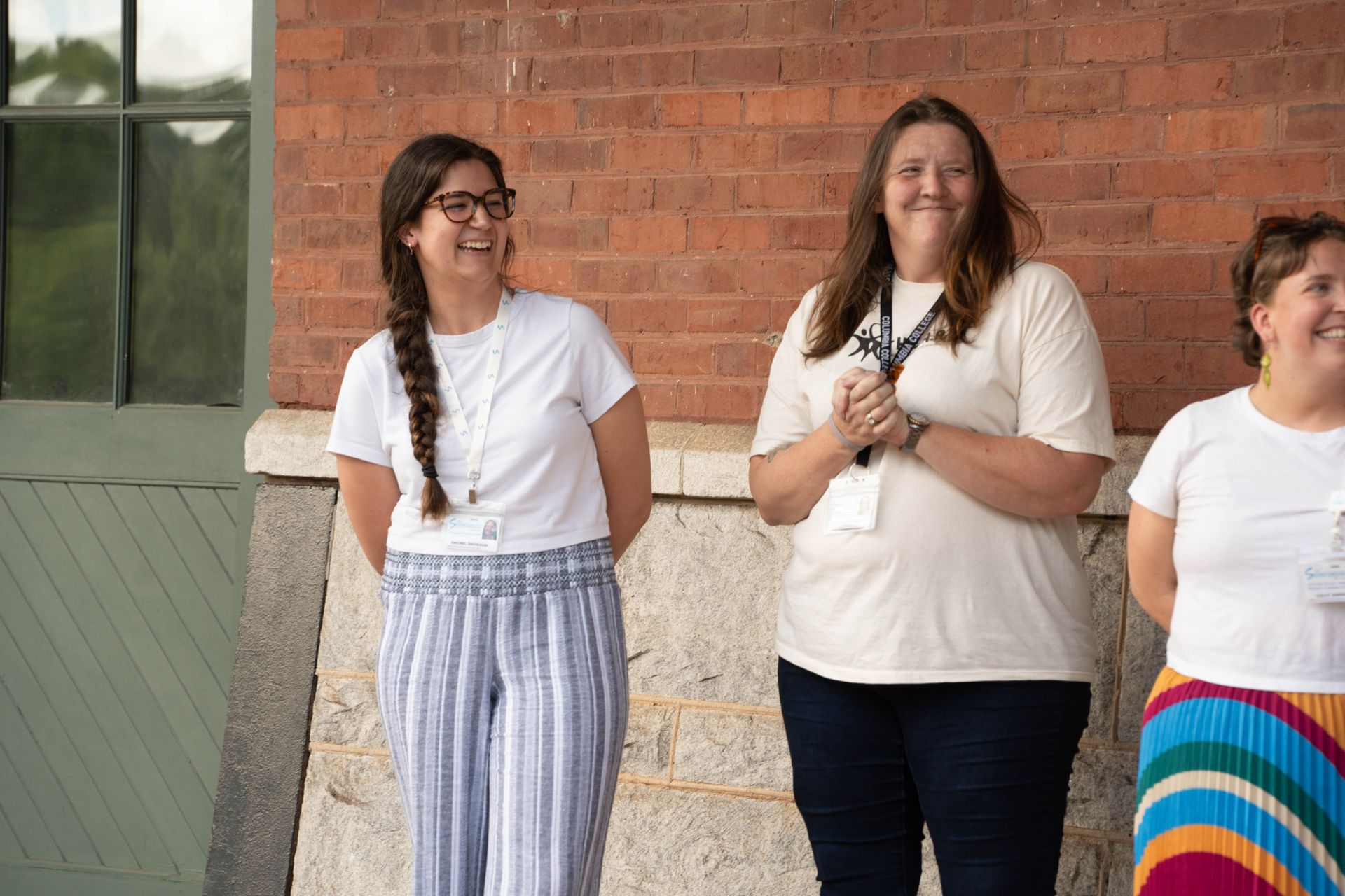 Three women smiling in front of a brick building. One wears a braid, glasses, and striped pants. The second has a name tag.
