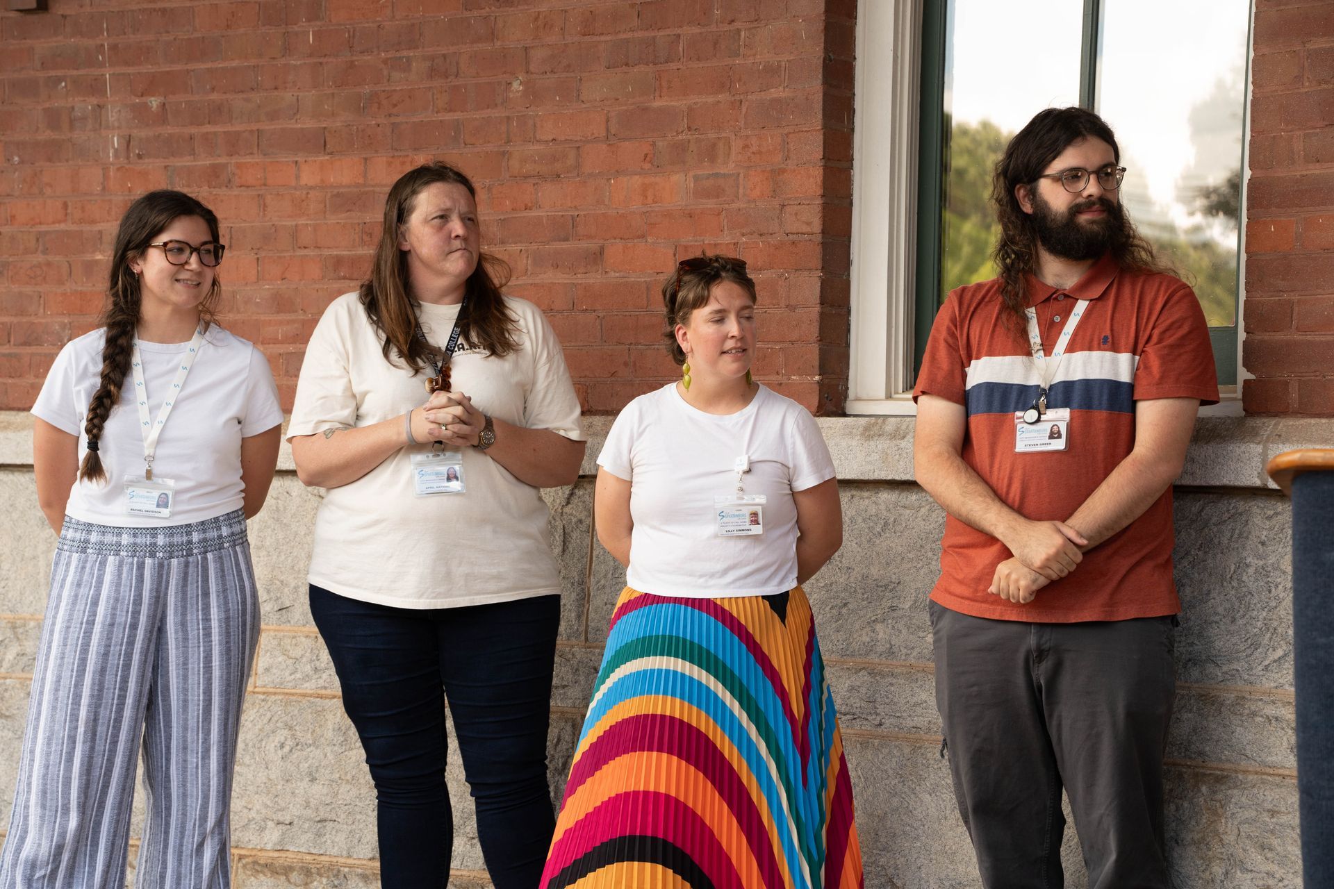 Four people standing in front of a brick wall. One in a rainbow skirt. All wearing lanyards, smiling.