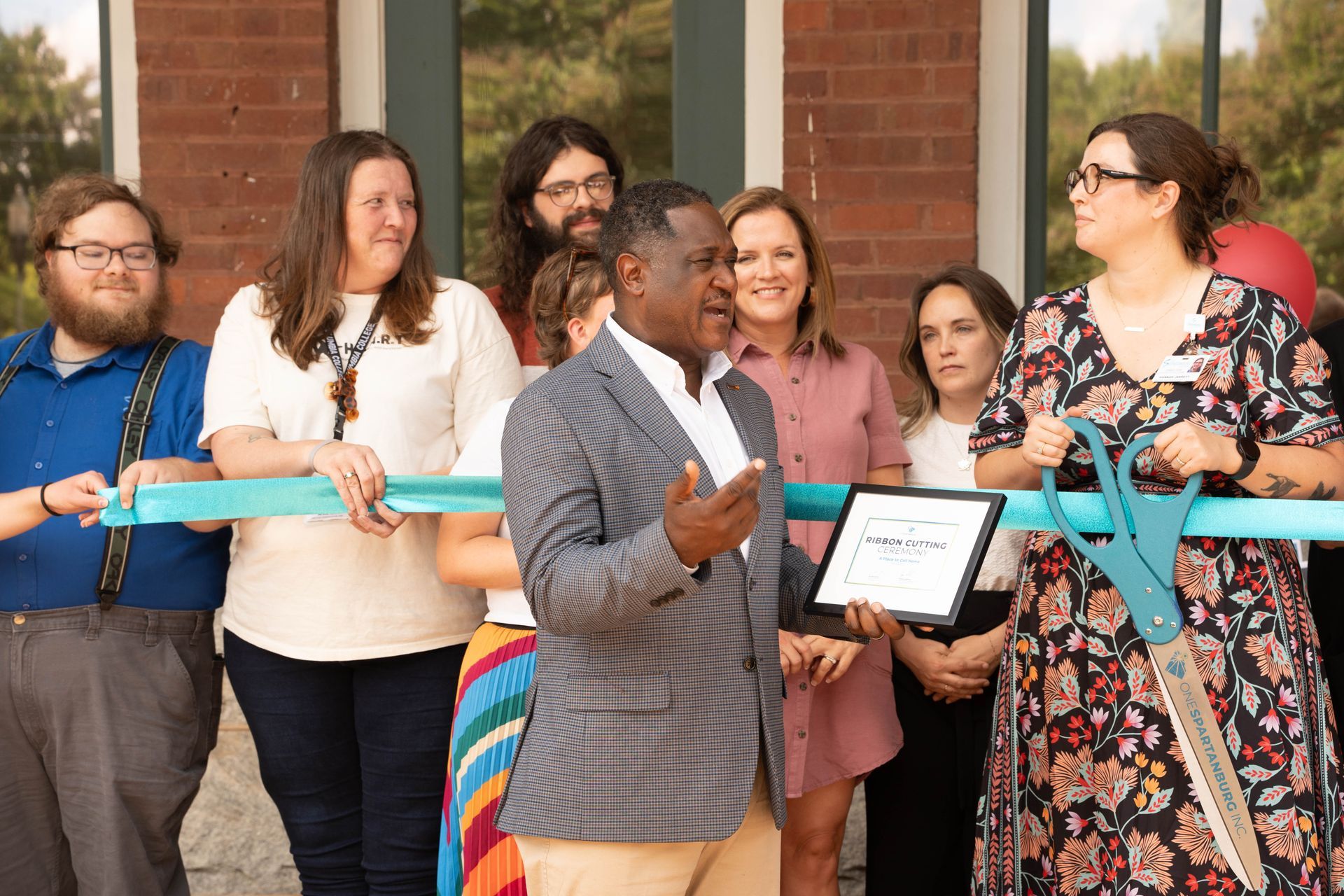 People cutting ribbon at a building opening. Man speaks, others smile, building in background.