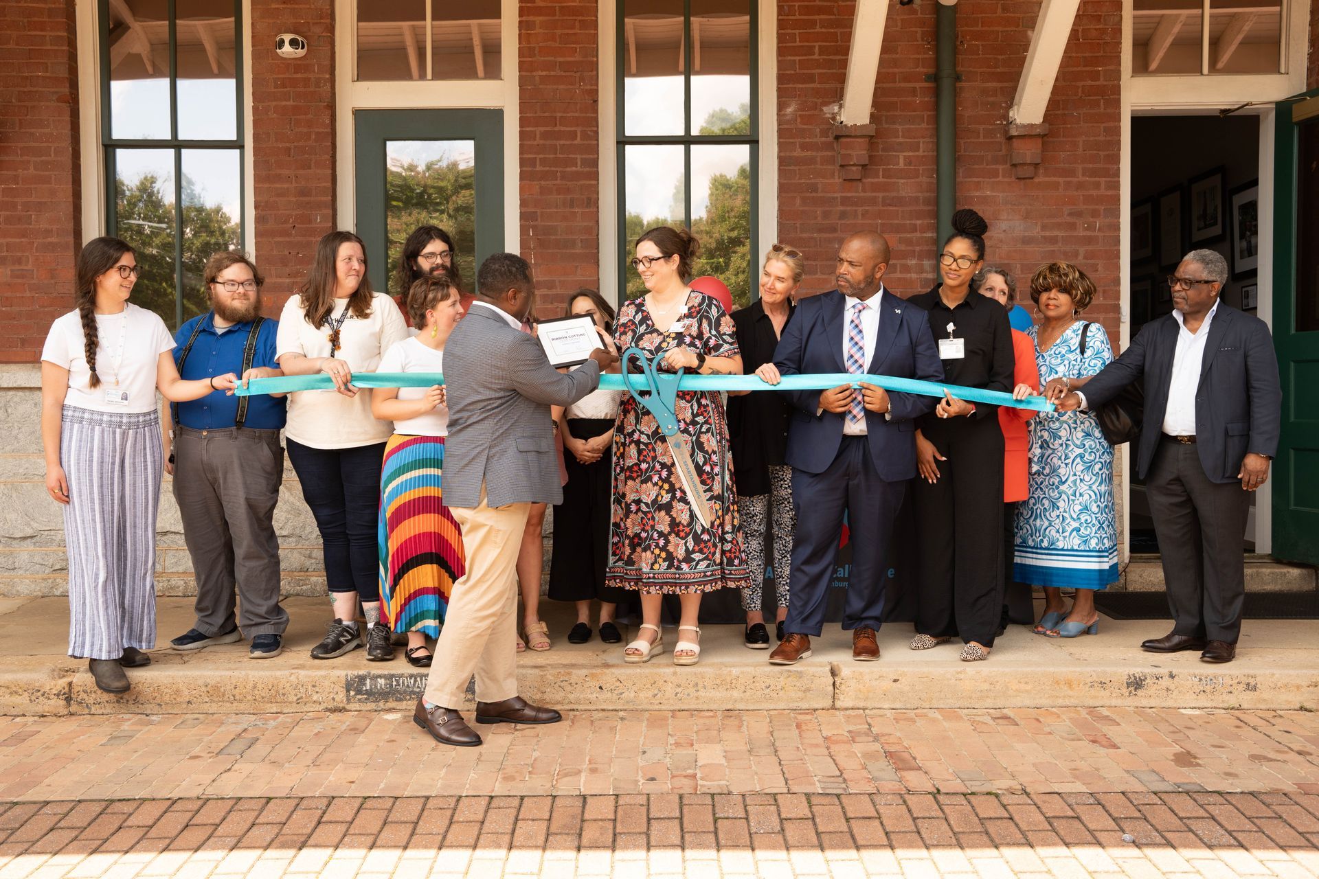 People cutting a ribbon in front of a brick building. Several people watch, smiling, on a sunny day.