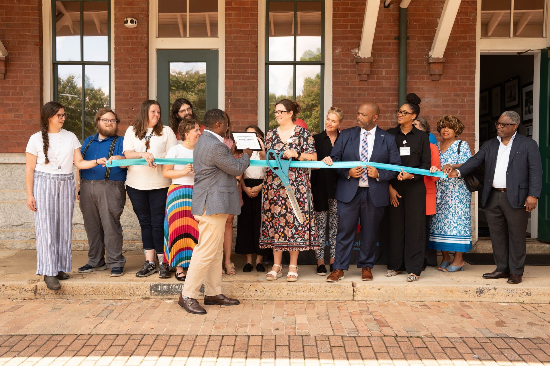 Group of people cutting a blue ribbon at a building's entrance. Brick exterior.