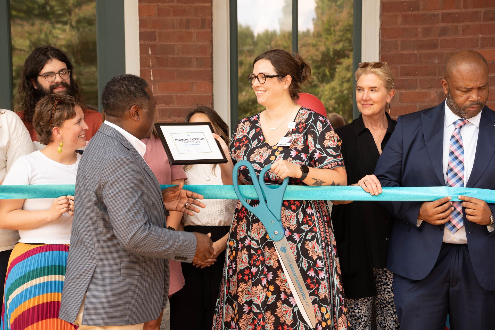 Group cuts teal ribbon with giant scissors. Brick building exterior.