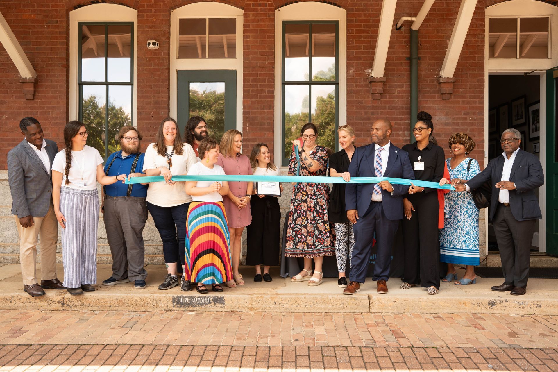 People cutting a ribbon at a building opening. Multi-ethnic group smiling, outdoors. Building is brick, with windows and a door.