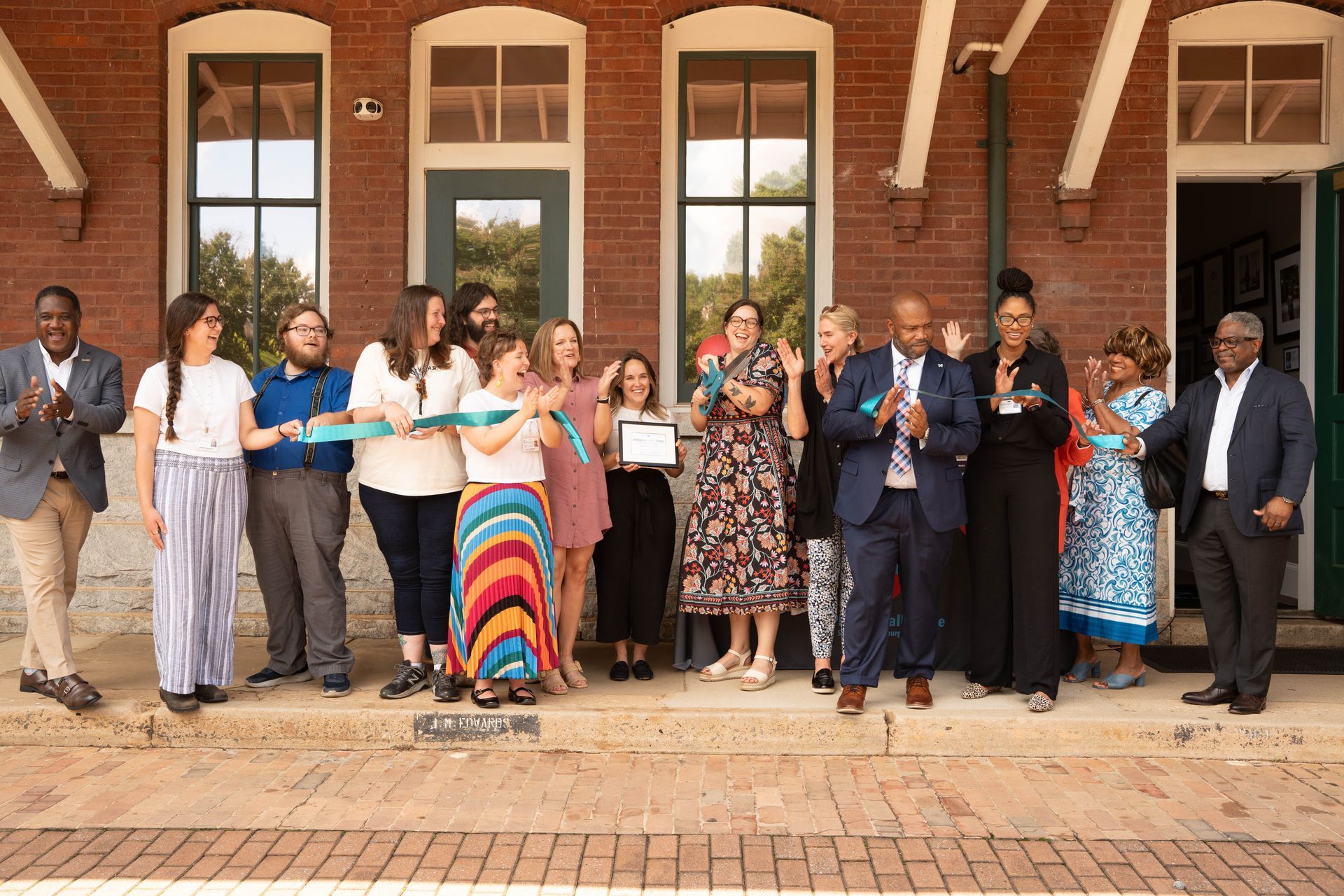 Group celebrates ribbon cutting outside a brick building. People clap, smile, and hold a certificate.