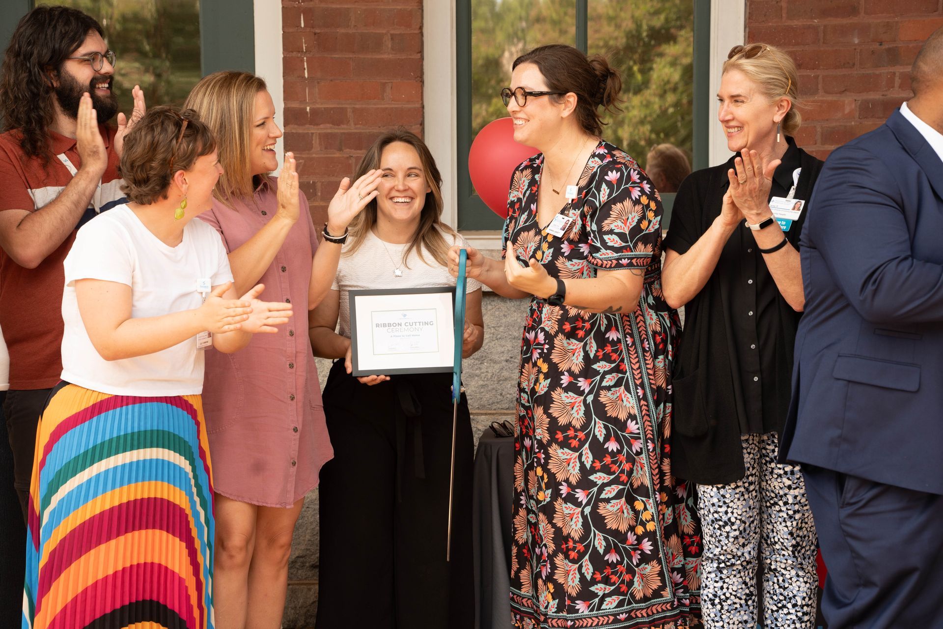 Group of people clapping, celebrating a woman holding a certificate outside a brick building.